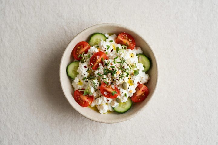 Overhead view of a small bowl of cottage cheese topped with cherry tomato halves, cucumber and fresh herbs